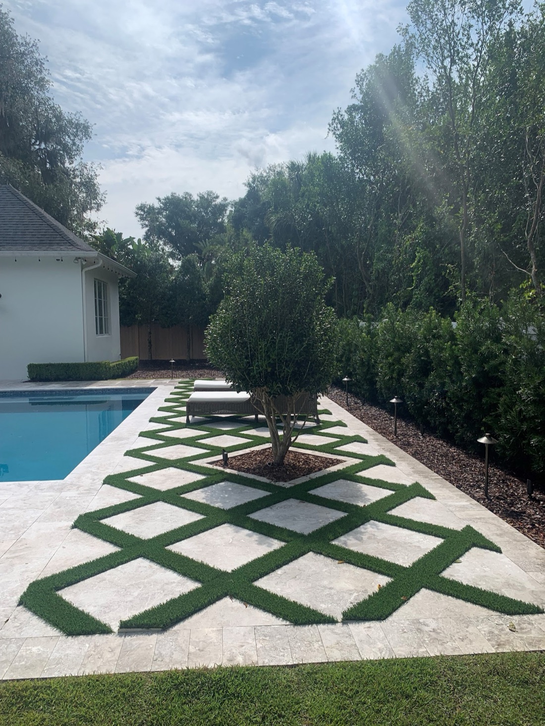 Travertine deck with diamond-pattern turf inlays and an ornamental tree, pool partially visible